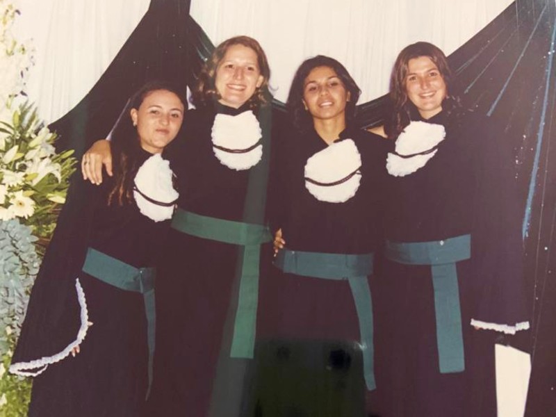Foto da formatura de Thais com mais três amigas, uma ao lado da outra. Elas usam beca preta, faixa verde, estão olhando para a frente e sorrindo
